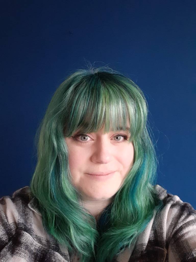 A white person with long, green hair smiling in front of a blue background.